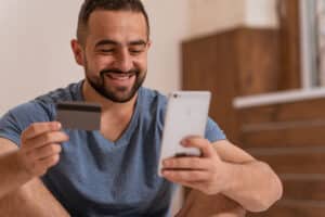 Happy man sitting on floor using credit card and phone.