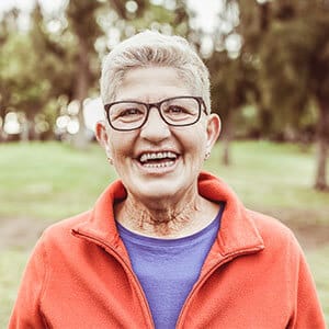 Happy older lady standing in park wearing light jacket and glasses.