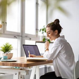 Woman sitting in well lit room at desk using laptop and writing in notebook.
