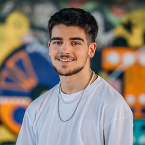 Young man standing in front of colorful mural in tshirt and necklace, smiling for photo.