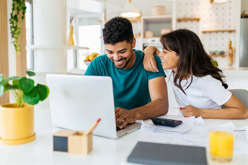 Young man and woman smiling and looking at one another while paying with credit card online at kitchen table.