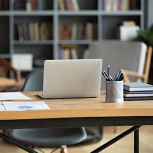 Desk in office with laptop and books resting on it and large bookshelf behind it.