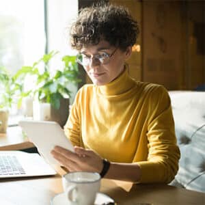 Woman sitting at desk with laptop and coffee cup resting on table while looking at tablet.