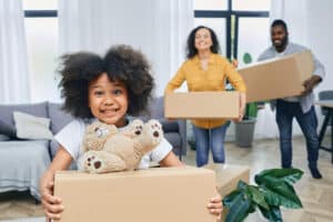 Man and woman with moving boxes walking behind little girl holding box with teddy bear resting on top.