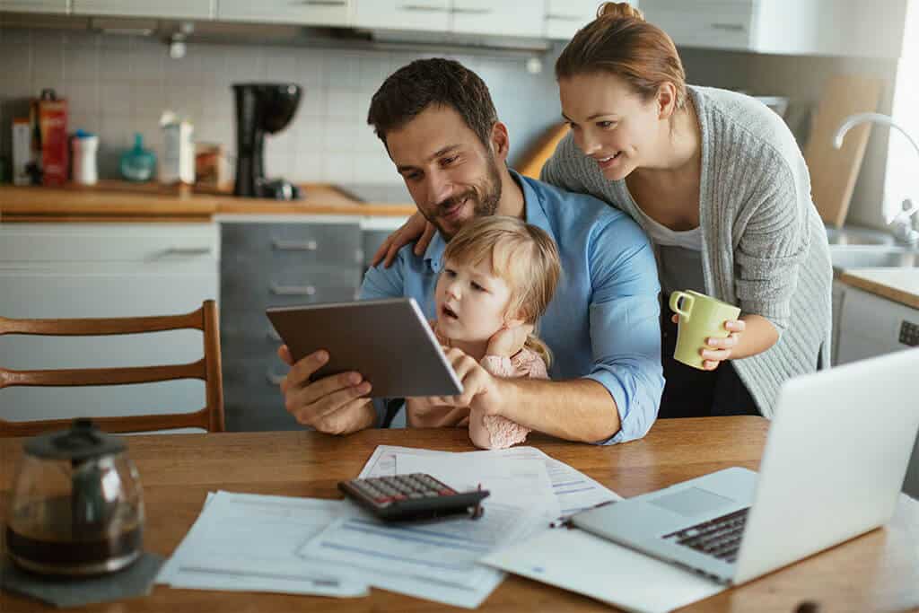 Family of three sitting together at kitchen table surrounded by documents, laptop, calculator and coffee pot, looking at tablet.