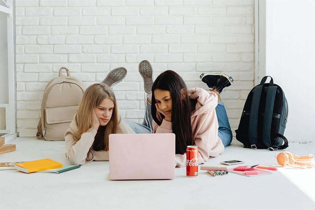 Two teenage girls laying on floor looking at laptop screen surrounded by bookbags and school supplies.