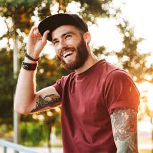 Hipster man with hat and tattoo sleeve on his arm smiling for photo with sun peering through trees behind him.