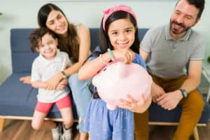 Mother, father and son sitting on couch looking at young daughter holding piggy bank and putting coin inside.