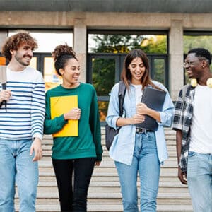 Group of students holding notebooks and carrying bookbags while smiling and walking in front of large building entrance.