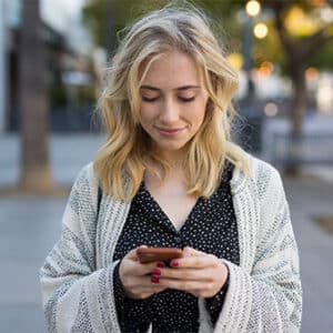 Woman standing on sidewalk looking down at her cell phone.