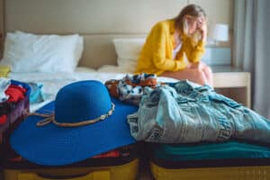 Overwhelmed woman with head resting on hand while sitting on the edge of bed with folded clothes resting on top of open suitcase.