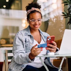 Happy lady sitting at desk with laptop in front of her looking at credit card in one hand and cell phone in another.