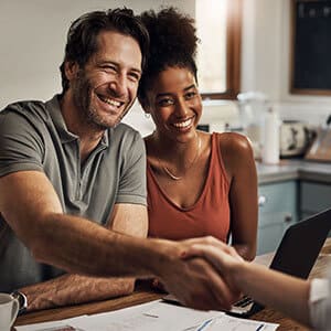 Multiracial couple sitting next to one another in kitchen while man stretches hand out to shake hand of person across the table.