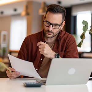 Man sitting at desk looking at document in hand with laptop sitting in front of him on table.