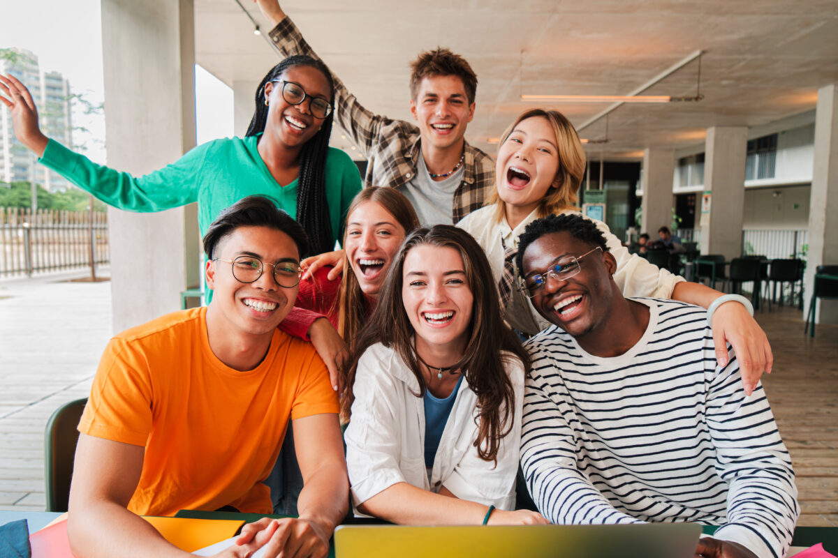 Diverse group of happy young adults smiling and posing together at an outdoor café with laptops, representing community, friendship, and teamwork.